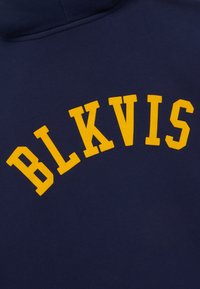 Tissu bleu marine avec des lettres jaunes audacieuses formant le mot « BLKVIS » en arc de cercle, probablement une partie d'un sweat-shirt ou d'un sweat à capuche.