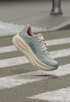 Zapatilla de running de color azul claro y beige con suela gruesa, flotando sobre un paso de peatones en una calle gris.