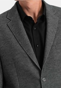 Szary teksturowany żakiet z subtelnym wzorem, klapami z wycięciem oraz pojedynczym zapięciem na guzik, noszony na czarnej koszuli z kołnierzykiem.