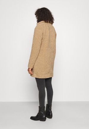 Beige nepbontjas met een zachte textuur, tot de knie en met een omslagkraag. Draagt over donkere jeans en zwarte enkellaarzen.