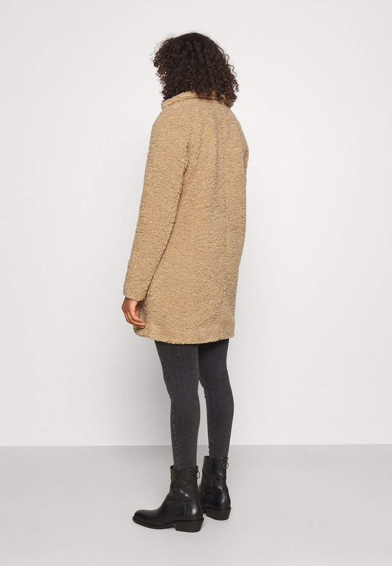 Beige kabát z umělého kožešina s měkkou texturou, do délky kolen, se sklápěcím límcem. Nosený přes tmavé džíny a černé kotníkové boty.