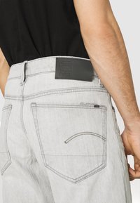 Jeans de mezclilla de color gris claro con parche de cuero negro de la marca en la cintura, costuras visibles en el bolsillo trasero, usados por una persona con camiseta negra.