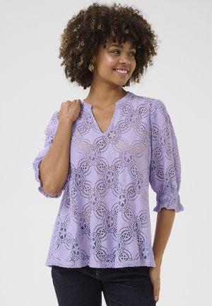 Blusa di pizzo lavanda con motivi floreali intagliati, maniche corte a sbuffo e scollatura a V. Texture morbida e design svasato con accenti arricciati.