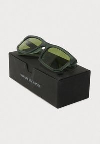 Žalios matinės plastikinės saulės akinių su stačiakampiais lęšiais, padėtos ant juodos dėžutės su užrašu "Armani Exchange."