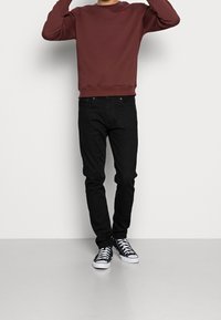 Sudadera burdeos con cuello redondo, vaqueros negros y zapatillas negras y blancas. La sudadera tiene un pequeño logo en el pecho.