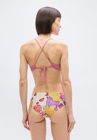 Γυναίκα με κοντά καστανά μαλλιά που φο ράει ροζ μπικίνι τοπ με σταυρωτούς ιμάντες και μπικίνι κάτω μέρος με λουλουδένιο σχέδιο και ψηλή μέση, γυρισμένη πλάτη σε λευκό φόντο.