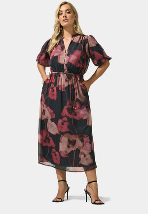 Kvinde iført en sort og pink blomsterprintet midikjole med pufærmer, et bindebånd ved taljen og sorte højhælede sandaler, står med hænderne på hofterne.
