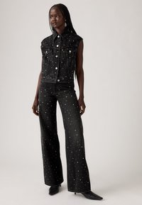 Zwarte denim mouwloze jas met zilveren studs, gecombineerd met high-waisted wijde jeans die ook zilveren studs hebben. Zwarte haklaarzen.