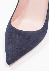 Marinblå mockapumps med spetsig tå, mjukt innerfoder och en slank silhuett, med matt textur och utan synliga detaljer i metall.