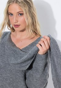 Grijze gebreide trui met een brede, gedrapeerde halslijn en geribbelde structuur. Model houdt een deel van de mouw vast, waardoor de zachtheid van de stof zichtbaar is.