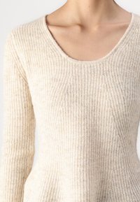 Kobieta nosząca sweter w kolorze kremowym, o prążkowanej fakturze i dekolcie w serek, eksponujący górną część klatki piersiowej i obojczyki.