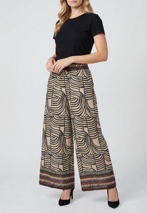 Vrouw die een zwart T-shirt met korte mouwen draagt, beige broek met wijde pijpen en zwarte geometrische patronen, en zwarte hoge hakken, die stil staat.