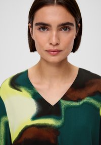 V-hals blouse in een groen, geel en bruin abstract patroon. Glad materiaal met een losse pasvorm, dat de levendige kleurenmix en de zachte textuur benadrukt.