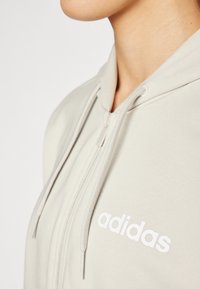 Svijetlosmeđa zip-up majica od mekane tkanine, s prednjim patentnim zatvaračem, vezicama i bijelim "adidas" logom na lijevoj strani prsnog dijela.