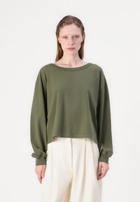 Oliva zöld hosszú ujjú crop top széles nyakkivágással és kényelmes szabással. Puha anyag, összegyűrt mandzsetták, fehér, laza szabású nadrággal párosítva.