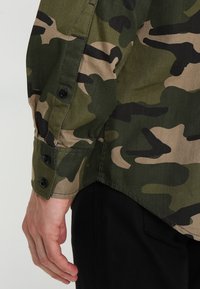 Camo shirt met een mix van donkergroene en tan patronen, gemaakt van textuurstof met zwarte knoppen en een gebogen zoom.