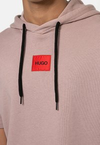 Primo piano di una felpa leggera color malva con cordini neri e una toppa quadrata rossa con l'etichetta "HUGO" sul petto.