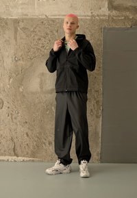 Μαύρο μπουφάν με φερμουάρ και κουκούλα, συνδυασμένο με χαλαρά γκρι παντελόνια. Λευκά αθλητικά παπούτσια με διακριτικές λεπτομέρειες. Λαμπερή υφή, casual σχέδιο.