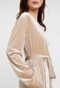 Roba di velluto beige chiaro con vita regolabile, maniche lunghe e design avvolgente. Il tessuto ha una texture morbida e liscia con una leggera lucentezza.