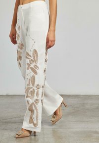 Pantaloni in lino bianchi con ricami floreali beige, caratterizzati da una vestibilità comoda e un design a gamba dritta. Scarpe con tacco completano l'outfit.