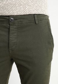 Primo piano di pantaloni verde oliva con bottone e passanti per cintura, indossati con un top in maglia beige testurizzato.