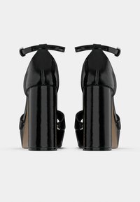 Μαύρα παπούτσια από πατέντα δέρμα με ψηλό τακούνι και χοντρό μπλοκ τακούνι, δετές στον αστράγαλο. Διαθέτουν ανοιχτό δάκτυλο και καμπυλωτή λεπτομέρεια στο πάνω μέρος.