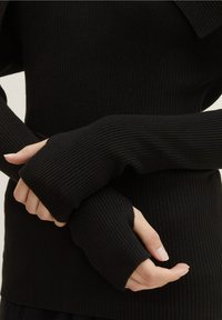 Mains croisées portant des manches longues noires côtelées avec des trous pour les pouces, laissant apparaître une peau beige et des ongles naturels sur un fond neutre.