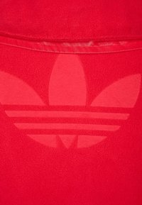 Červená mikina s kapucí, na které je tónový logo Adidas s třemi listovými tvary nad třemi horizontálními pruhy na strukturálním povrchu.