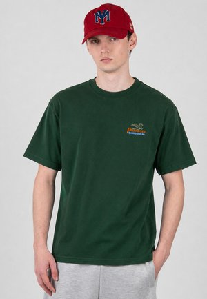Ung mand iført en mørkegrøn T-shirt med logoet "Prohibited Sporting Goods Inc." og en rød kasket med blå "NY" bogstaver, stående foran en grå baggrund.