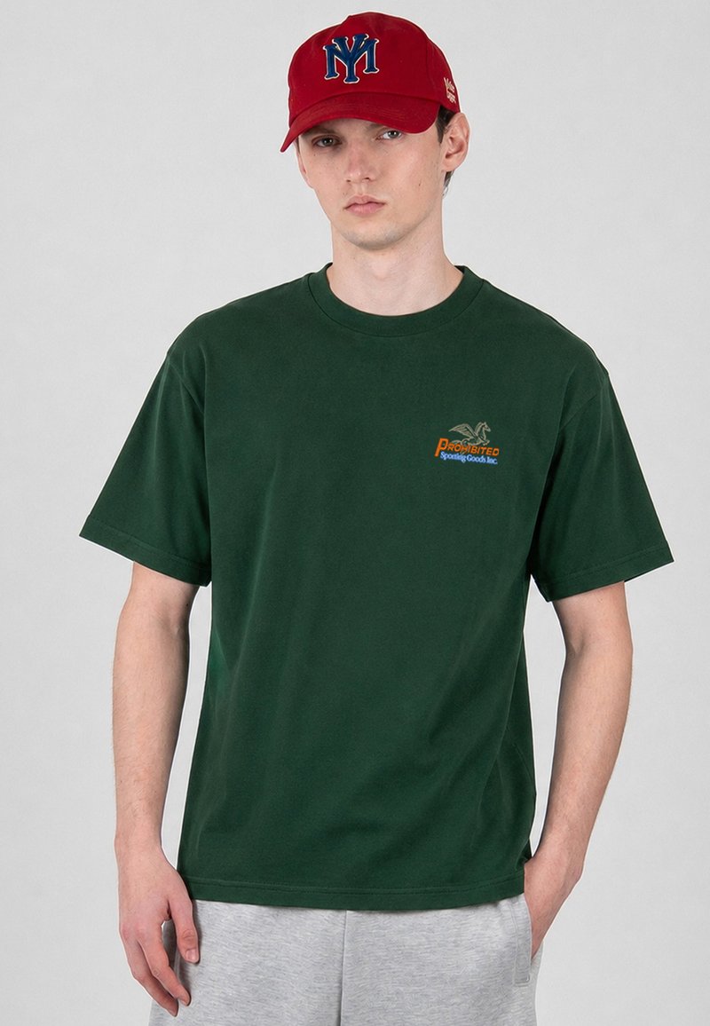 Νεαρός άνδρας που φοράει σκούρο πράσινο T-shirt με το λογότυπο "Prohibited Sporting Goods Inc." και κόκκινο καπέλο με μπλε γράμματα "NY", στέκεται μπροστά σε γκρι φόντο.