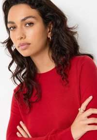 Rød strikket sweater med rund hals og lange ærmer. Modellen har mørkt, bølget hår og bærer hoops øreringe. Glat tekstur.