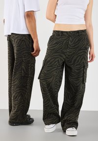 Cargo pants in olive green with black zebra stripes, featuring side pockets and a relaxed fit. Textured fabric with a durable design.
