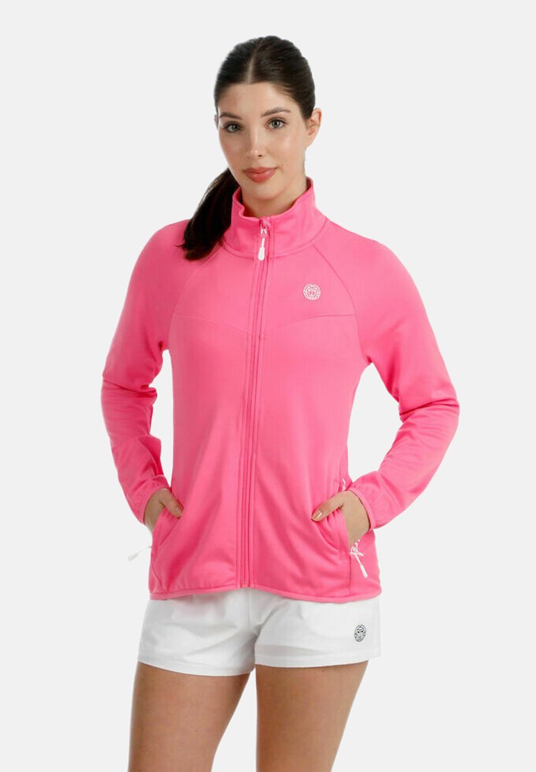 Frau mit dunklen Haaren, die eine leuchtend pinke Jacke mit Reißverschluss und Taschen sowie weiße Shorts trägt, vor einem schlichten hellen Hintergrund.