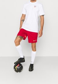 T-shirt Nike blanche avec un petit logo, shorts rouges avec un accent orange, chaussettes blanches et chaussures de football Nike noires sur un ballon de football noir.