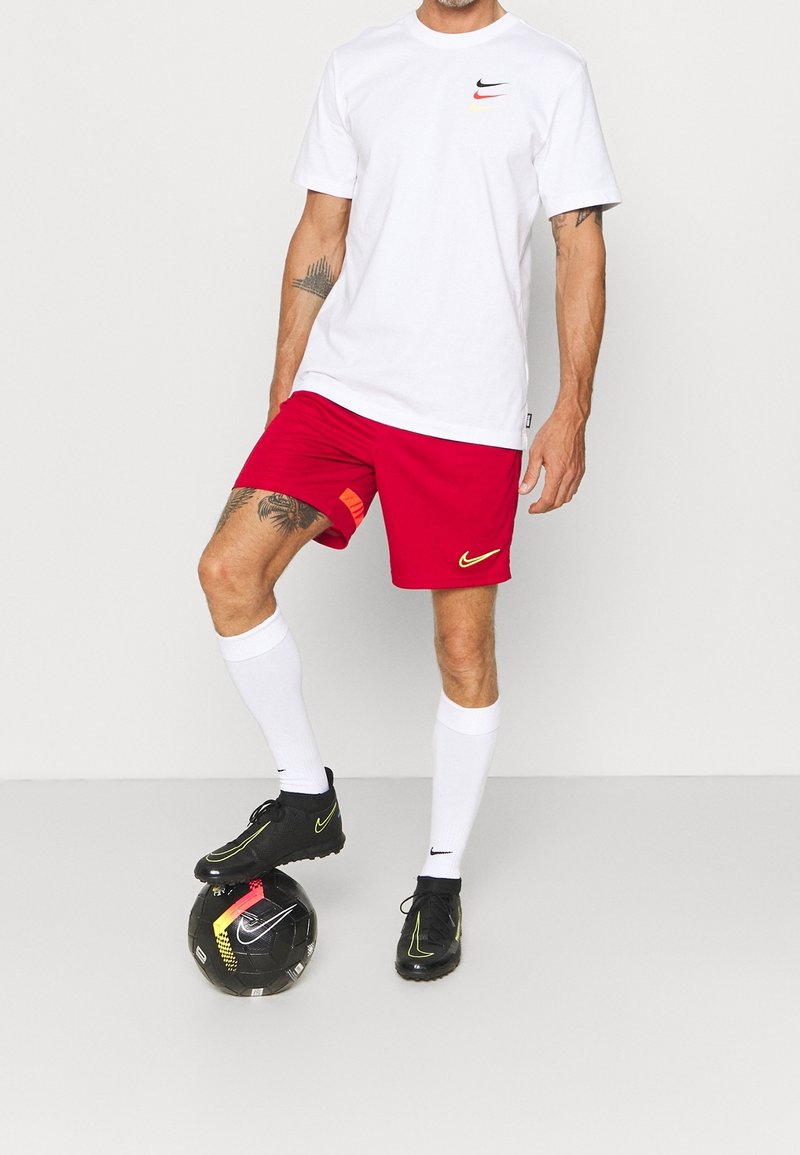 T-shirt Nike blanche avec un petit logo, shorts rouges avec un accent orange, chaussettes blanches et chaussures de football Nike noires sur un ballon de football noir.