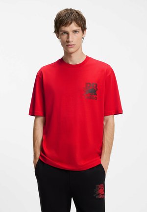Giovane uomo con capelli castani corti indossa una maglietta rossa e pantaloni neri, entrambi con piccoli loghi neri e rossi, in piedi davanti a uno sfondo bianco.