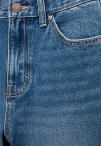 Blå denimjeans med en synlig framficka, metallknappstängning, kontrasterande orange sömmar och en något blek textur.