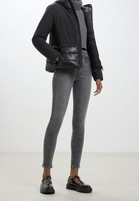 Donna che indossa un piumino nero, un maglione a collo alto grigio a coste, jeans skinny grigi e scarpe con tacco in vernice nera, in piedi all'interno.