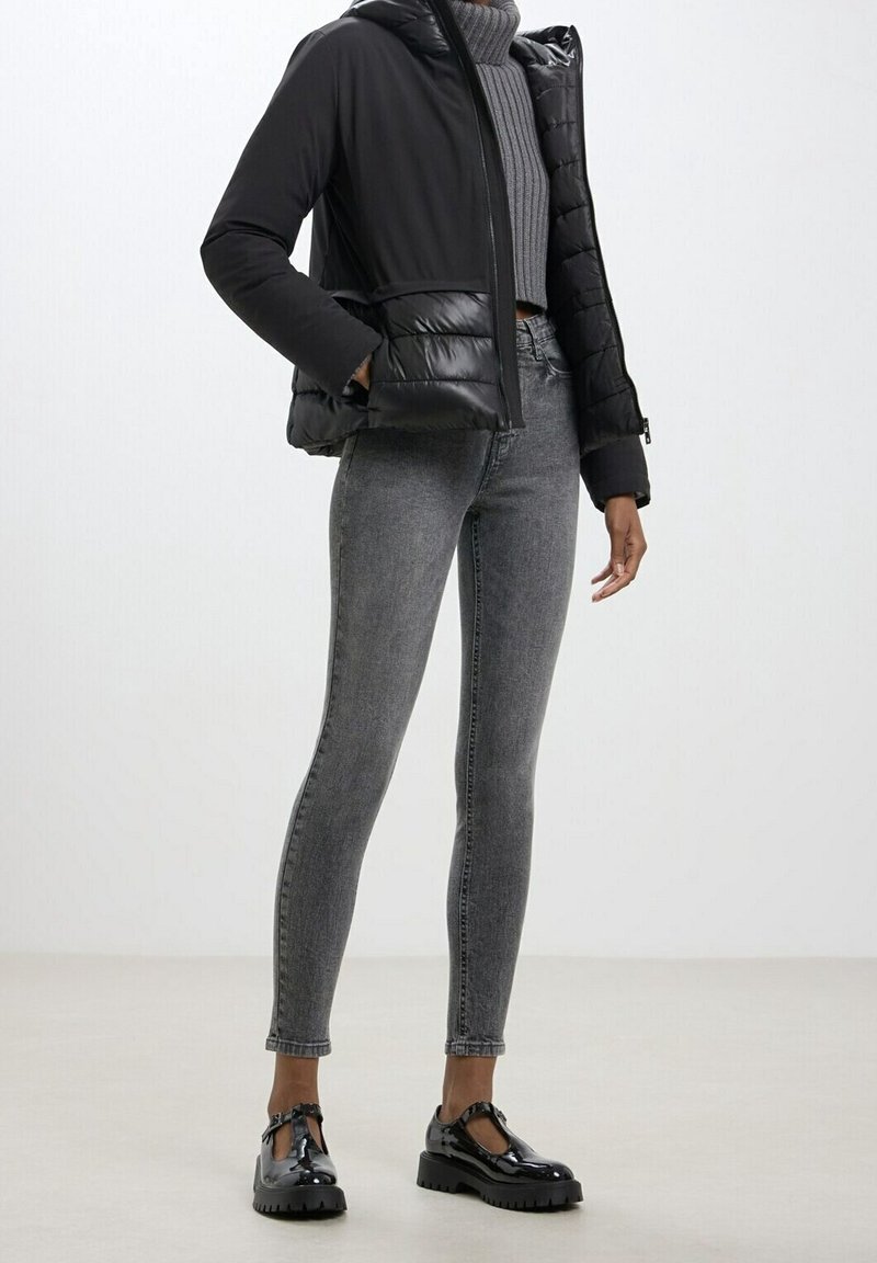 Donna che indossa un piumino nero, un maglione a collo alto grigio a coste, jeans skinny grigi e scarpe con tacco in vernice nera, in piedi all'interno.