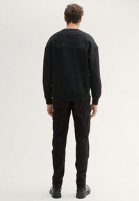 Svart sweatshirt med texturerad yta, med en stor präglad logotyp "TOM TAILOR" på baksidan, i kombination med svarta cargobyxor och kängor.