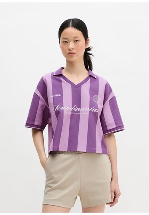 Γυναίκα που φοράει μωβ και λεβαντίνι ριγέ πόλο πουκάμισο με το κείμενο "Scandinavia Football Community" και μπεζ σορτς, στέκεται μπροστά σε λευκό φόντο.