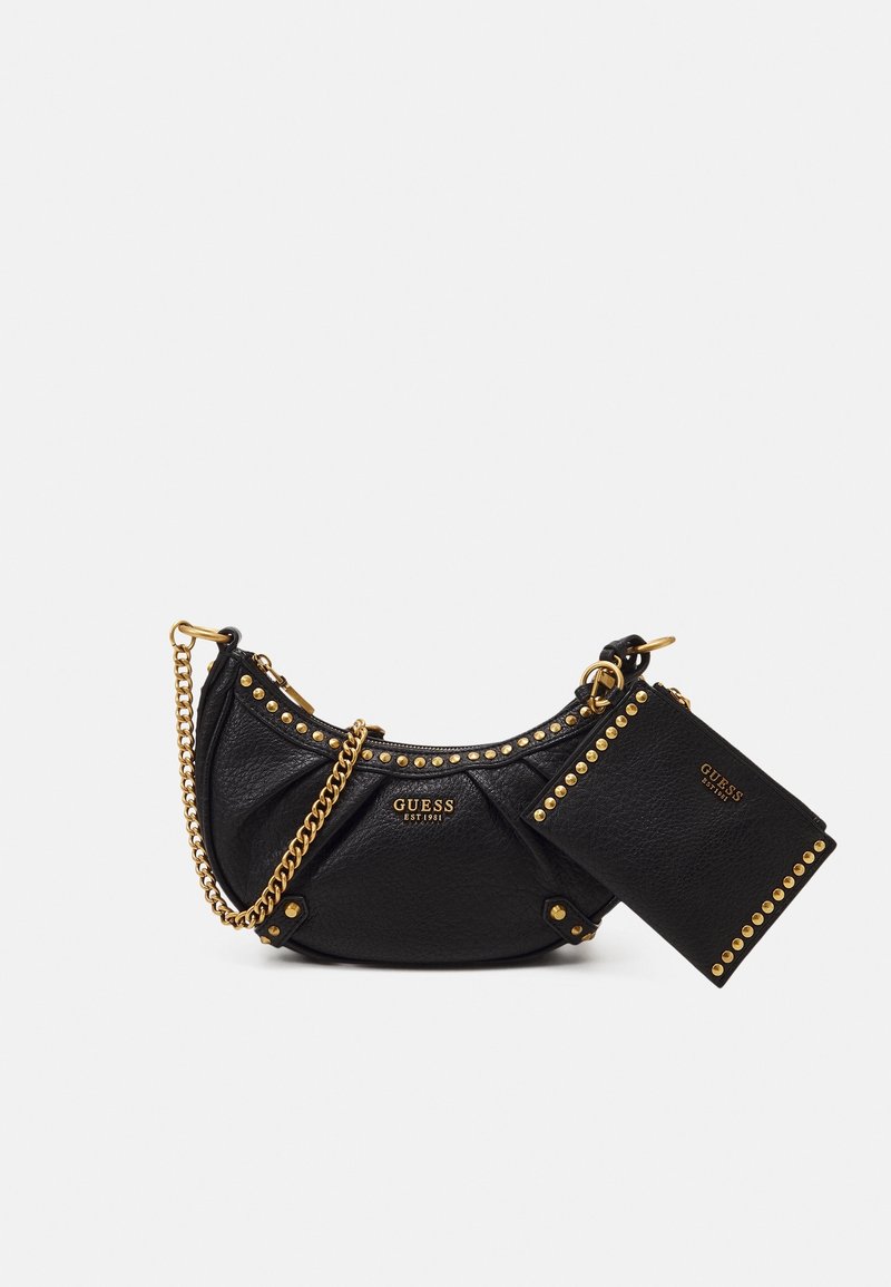 Czarna torebka marki Guess w kształcie półksiężyca z złotymi ćwiekami, łańcuszkowym paskiem oraz dopasowaną małą saszetką przymocowaną złotym kółkiem na białym tle.