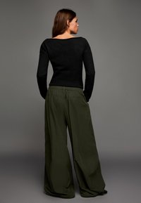 Fekete kötött pulóver széles nyakkivágással, laza fazonú, sima, könnyű anyagból készült olívazöld nadrággal párosítva.