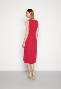 Vestido justo sem mangas em rosa brilhante, até ao joelho, com um discreto fecho nas costas e fendas laterais, feito de tecido suave. Um par de sapatos de salto ligeiramente bege.