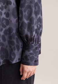 Dunkel gemusterte Bluse mit einem subtilen Leopardenprint. Satinähnliches Material mit gerafften Ärmeln und geknöpften Manschetten. Dunkle Knöpfe betonen die Manschetten.