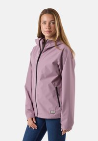 Młoda kobieta w liliowej wodoodpornej kurtce z kapturem, stojąca z rękami rozluźnionymi, na jednolitym jasnym tle.