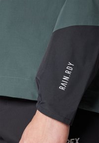 Vanntett jakke med grønn kropp og svarte ermer. Ermet har teksten "RAIN.RDY" i hvitt. Glatt og funksjonell design.