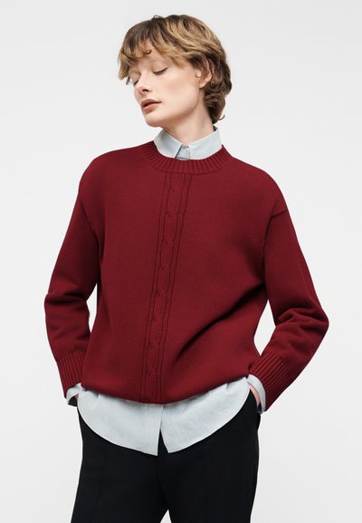 Άτομο που φοράει βυσσινί πουλόβερ πλεκτό με κοτσίδες πάνω από ένα ανοιχτό μπλε ριγέ πουκάμισο με κουμπιά, μαύρο παντελόνι, με τα χέρια στις τσέπες, κοιτώντας προς τα κάτω αριστερά.