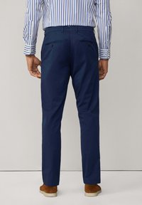 Pantaloni blu navy con una vestibilità su misura, dotati di due tasche posteriori e una texture del tessuto liscia. Indossati con una camicia a righe e scarpe marroni.