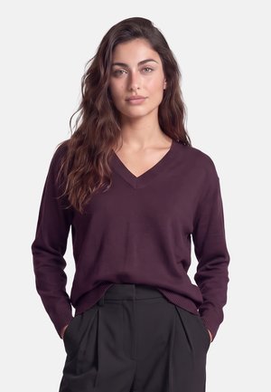Junge Frau mit langem, welligem Haar, die einen dunkelvioletten V-Ausschnitt-Pullover und schwarze plissierte Hosen trägt, Hände in den Taschen, neutraler Hintergrund.